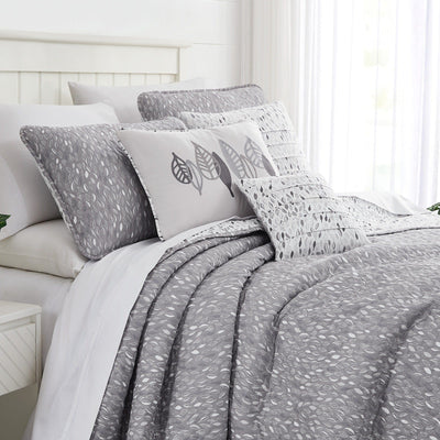 Angled view of Botanical Leaves quilt set showcasing gray leaf print quilt and matching shams on a bed with white paneled headboard