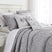 Angled view of Botanical Leaves quilt set showcasing gray leaf print quilt and matching shams on a bed with white paneled headboard