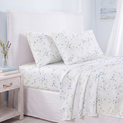 White bedding set with a delicate blue and green floral print, styled on a bed with a white upholstered headboard, a light wood nightstand, and a soft window curtain. #color_soft-bloom-blue